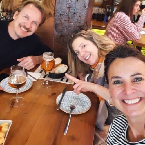 Clientes da Thais almoçando junto com ela em um dos roteiros de viagem. Eles bebem chopp e estão sorrindo.